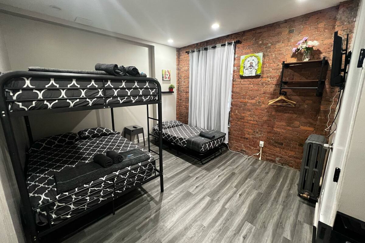 Una habitación con literas, una mesita, un enchufe, un radiador, un muro de ladrillo y una ventana con cortinas.