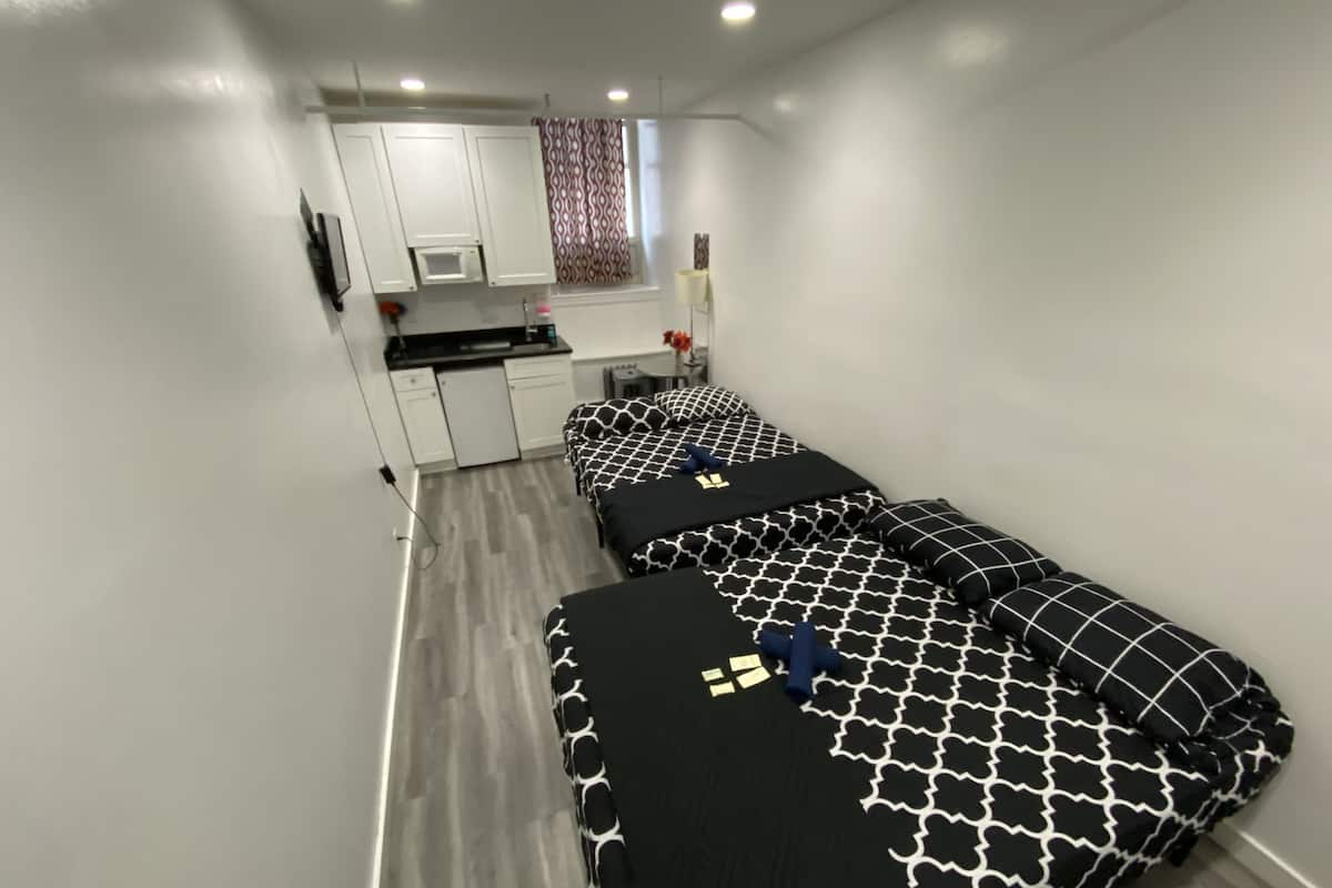 Una habitación funcional con dos camas, kitchenette y baño pequeño.