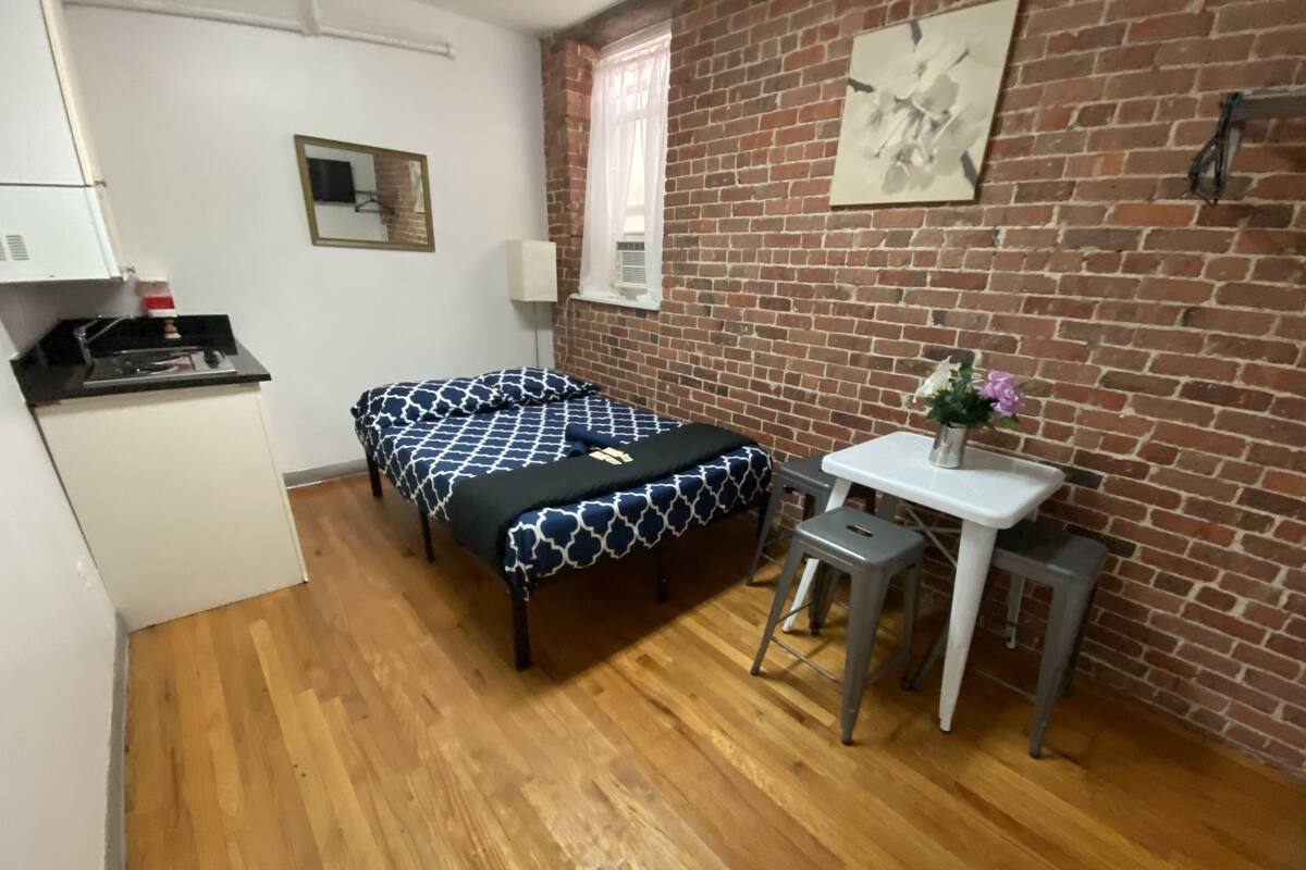 Una habitación compacta con dos camas, una kitchenette pequeña, una mesa blanca con pileta y una pared de ladrillo.