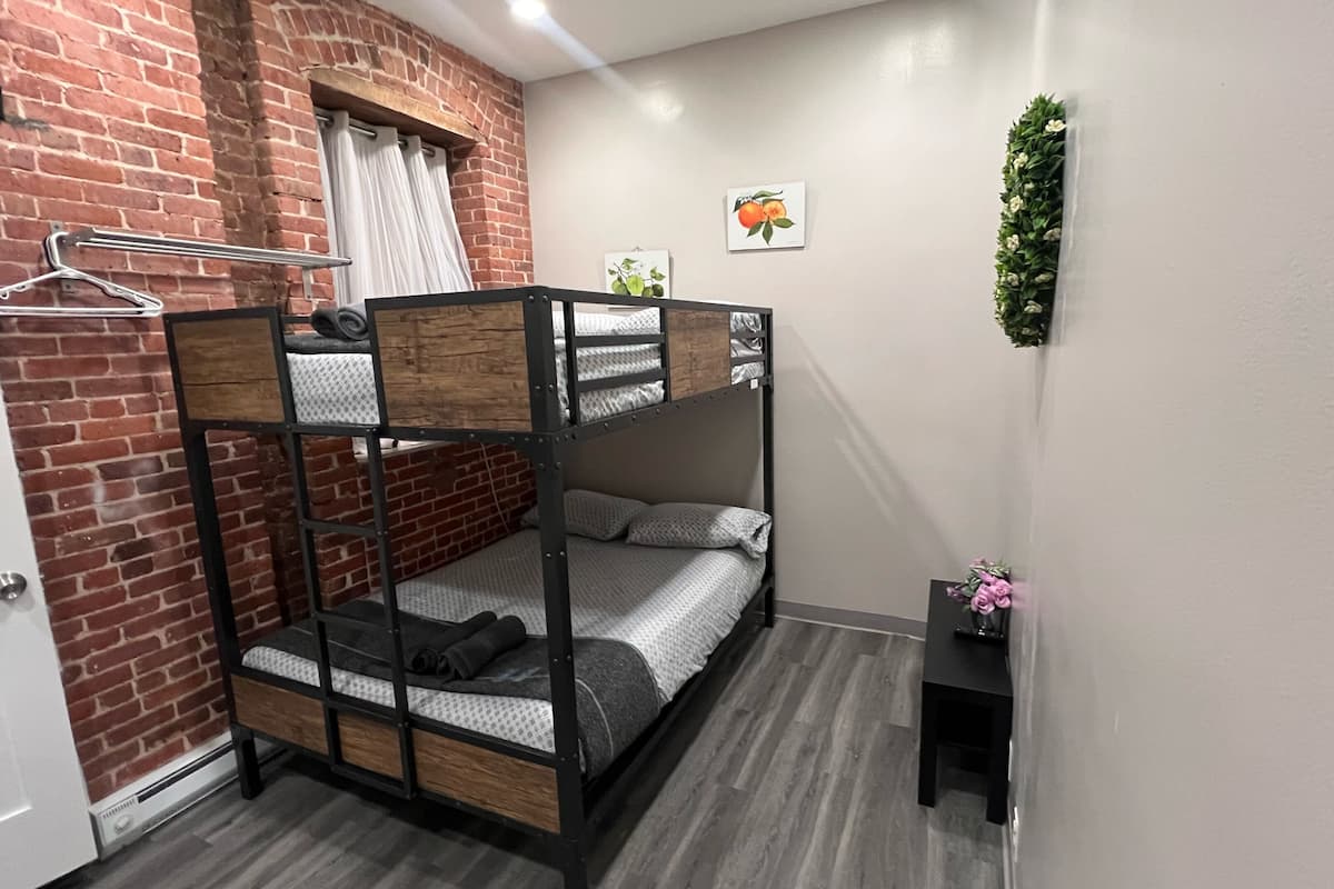 Habitación con literas, pared de ladrillo, puerta blanca y una pequeña mesa con un florero.