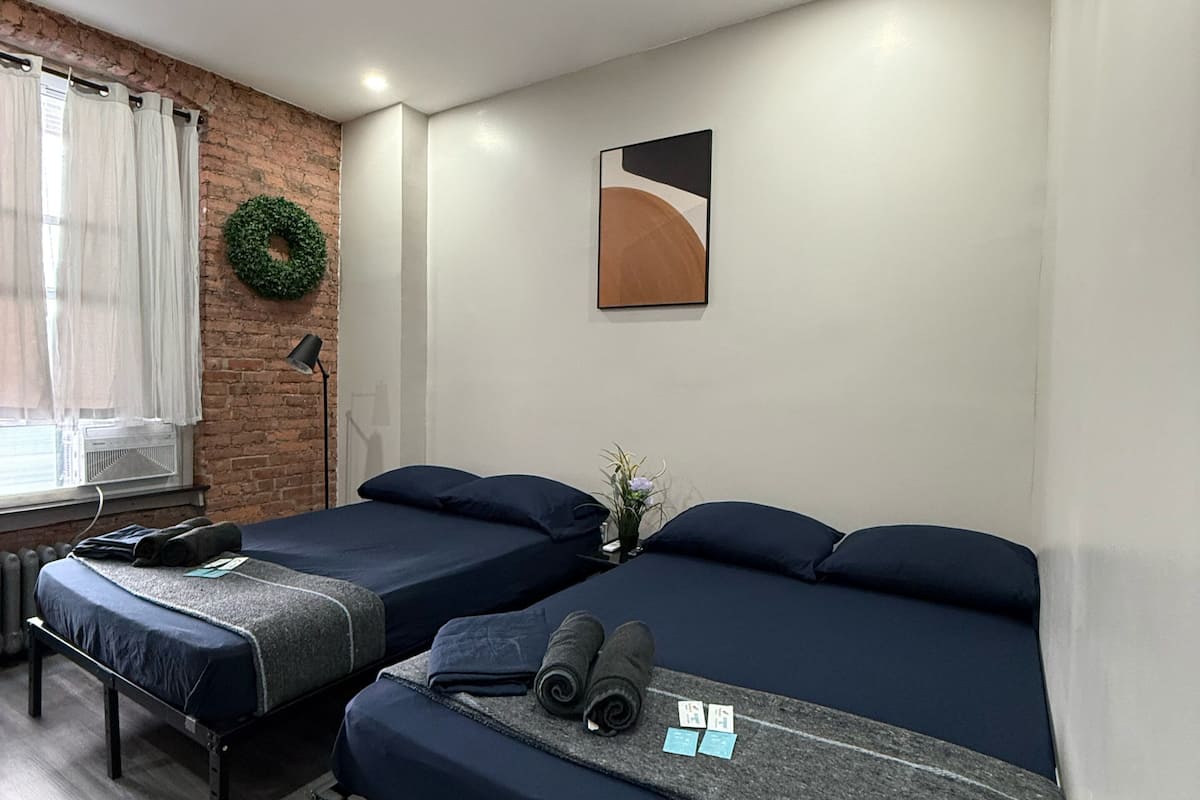 Una habitación con dos camas, una pared de ladrillo, una guirnalda verde, un cuadro y una mesita con una lámpara.