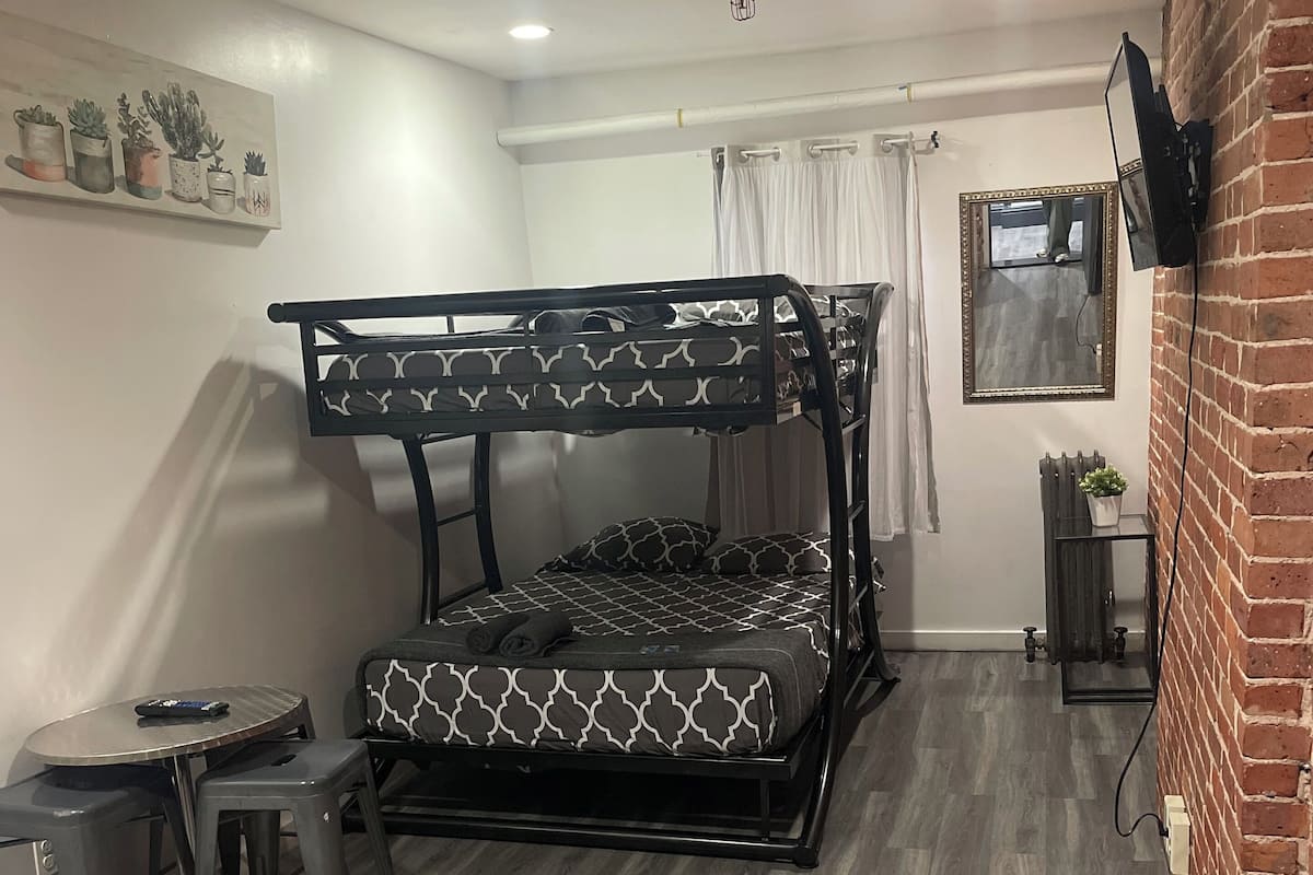 Una cama individual con un colchón estampado, una mesita con un control remoto, un taburete, un televisor fijado en una pared de ladrillo y una planta en maceta.