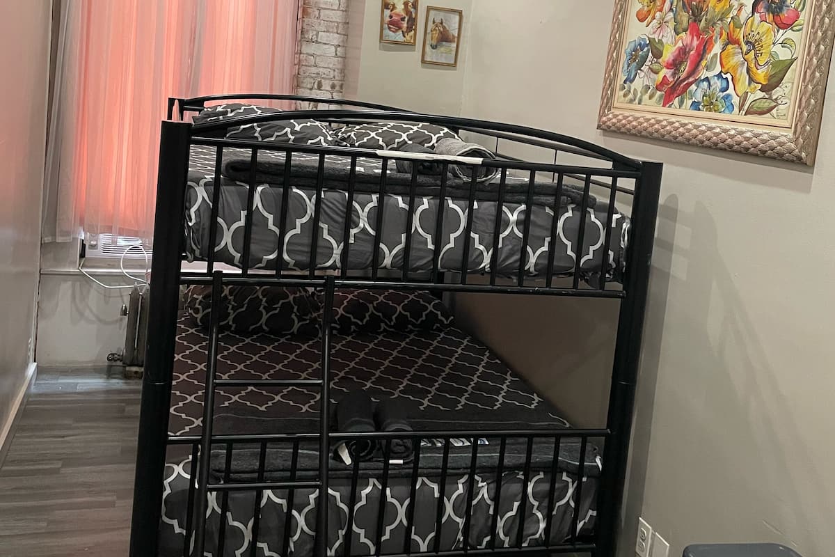 Una cama litera con un colchón estampado, una pared de piedra, una ventana con cortinas rosadas y un cuadro floral enmarcado en la pared.
