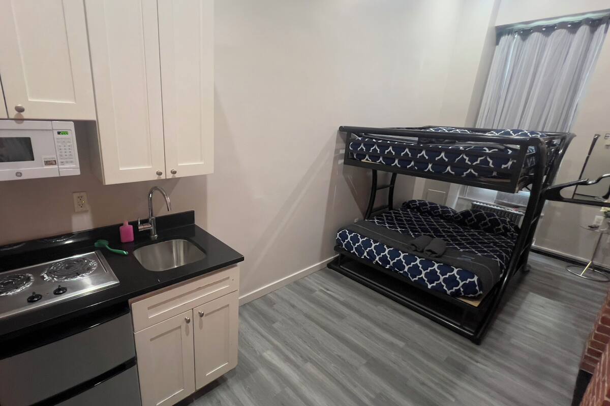 Cocina compacta con microondas, fregadero y estufa. También se observa una litera con ropa de cama estampada.