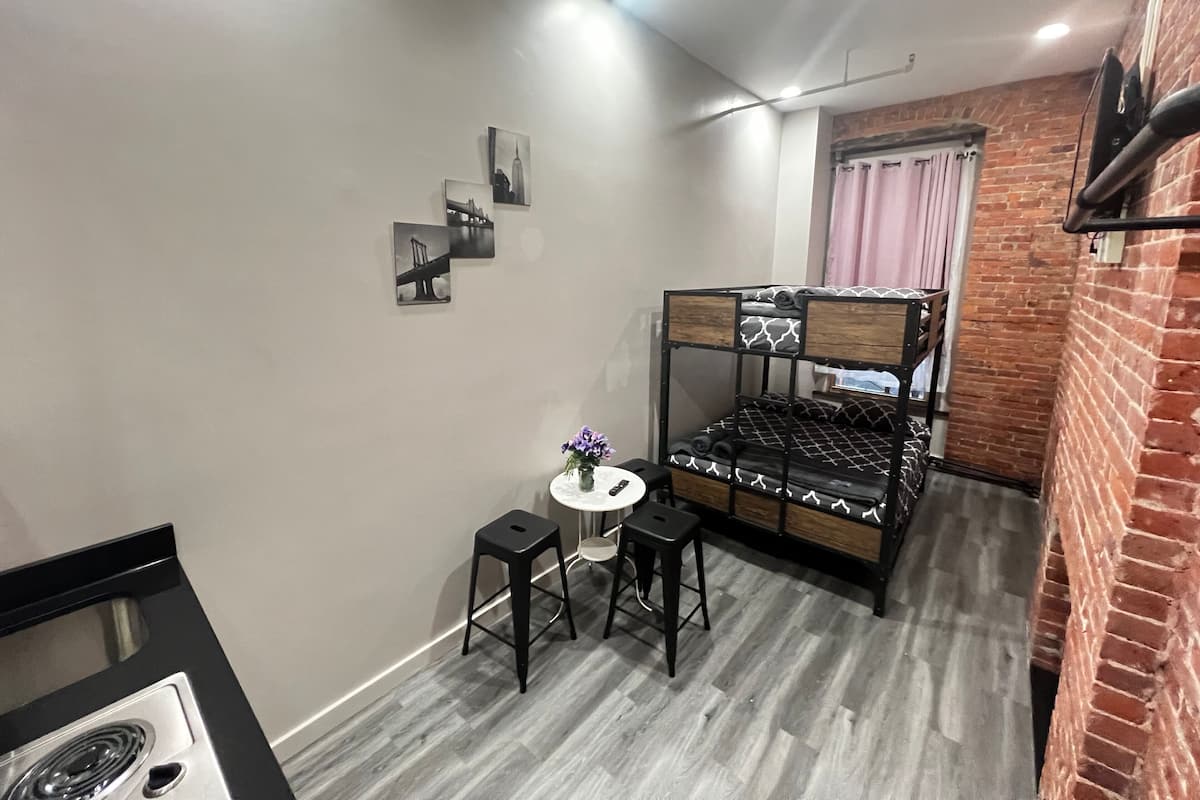 Una habitación compacta con literas, una mesita pequeña con dos taburetes, una cama individual con colcha estampada y una estantería fijada a la pared.