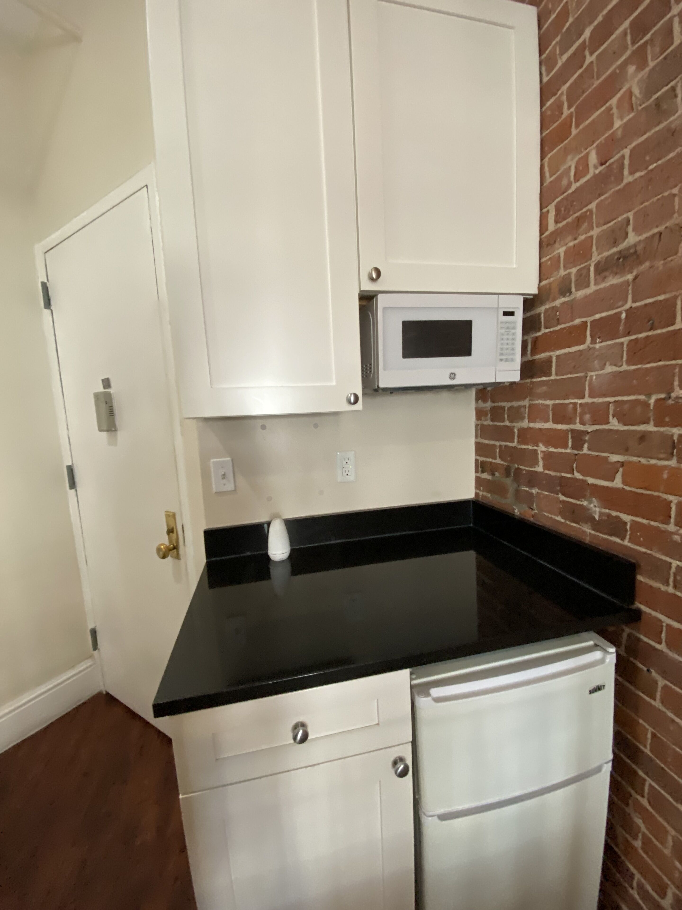 Cocina compacta con armarios blancos, encimera negra, microondas integrado y refrigerador de acero inoxidable.