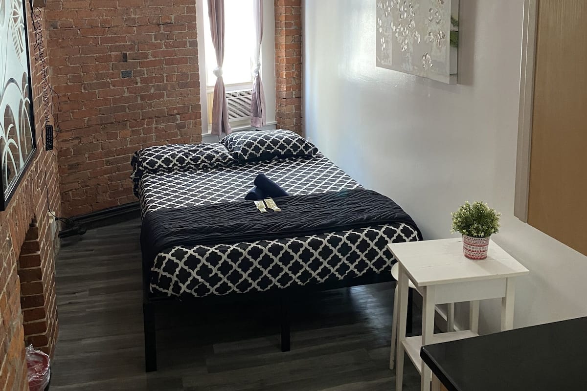Una habitación pequeña y moderna con pared de ladrillo, cama con ropa de cama estampada, una mesa pequeña con planta en maceta y ventana con cortinas.