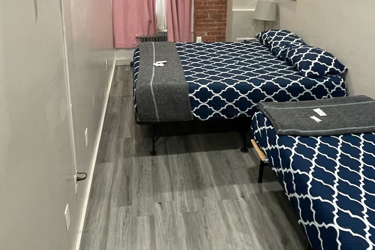 Una habitación pequeña con dos camas, un televisor montado en la pared, un muro de ladrillo y una ventana con cortinas rosadas.