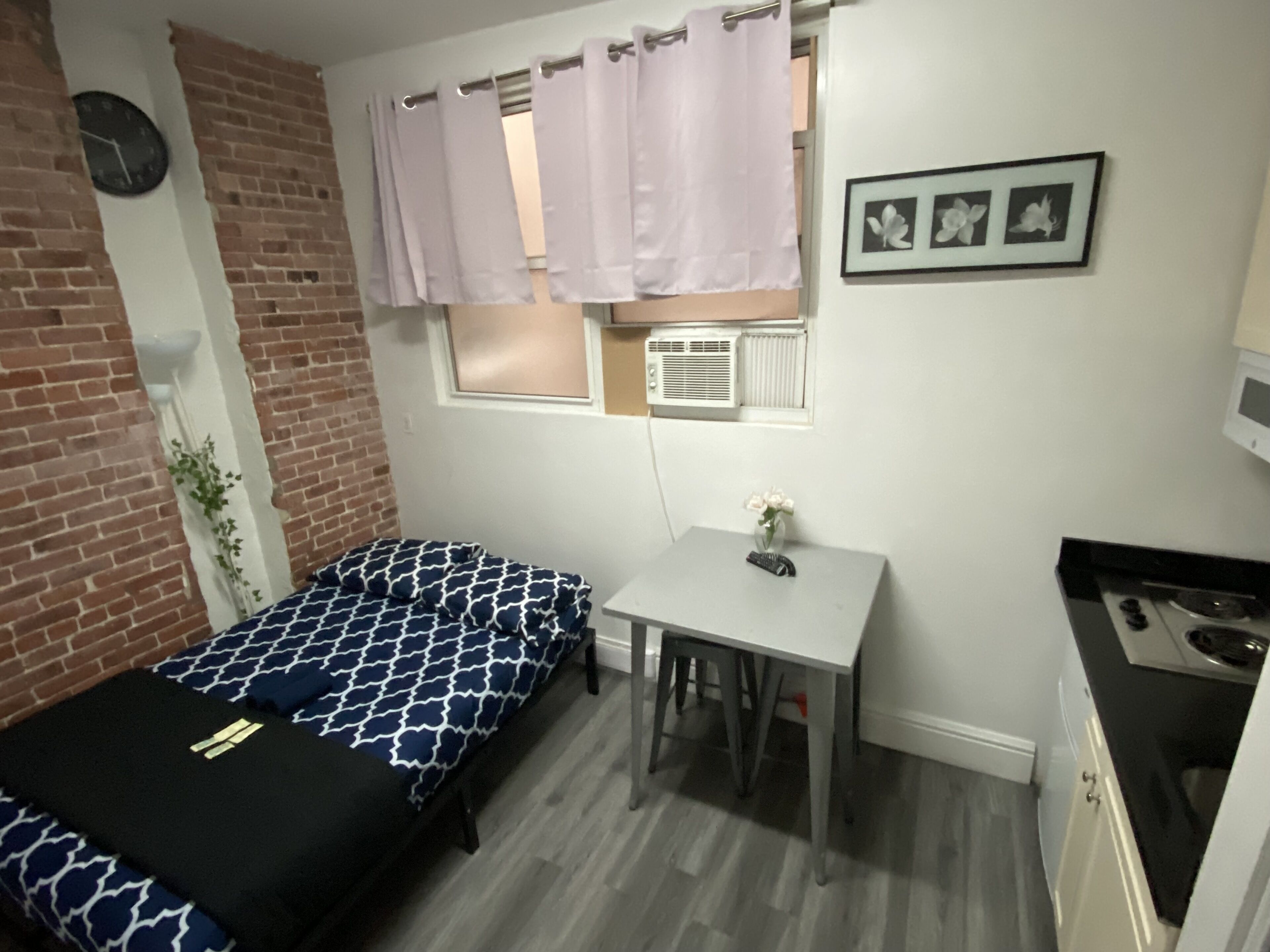 Une petite pièce comprenant un lit, une petite table, une fenêtre avec des rideaux, une horloge et un tableau encadré accroché au mur.
