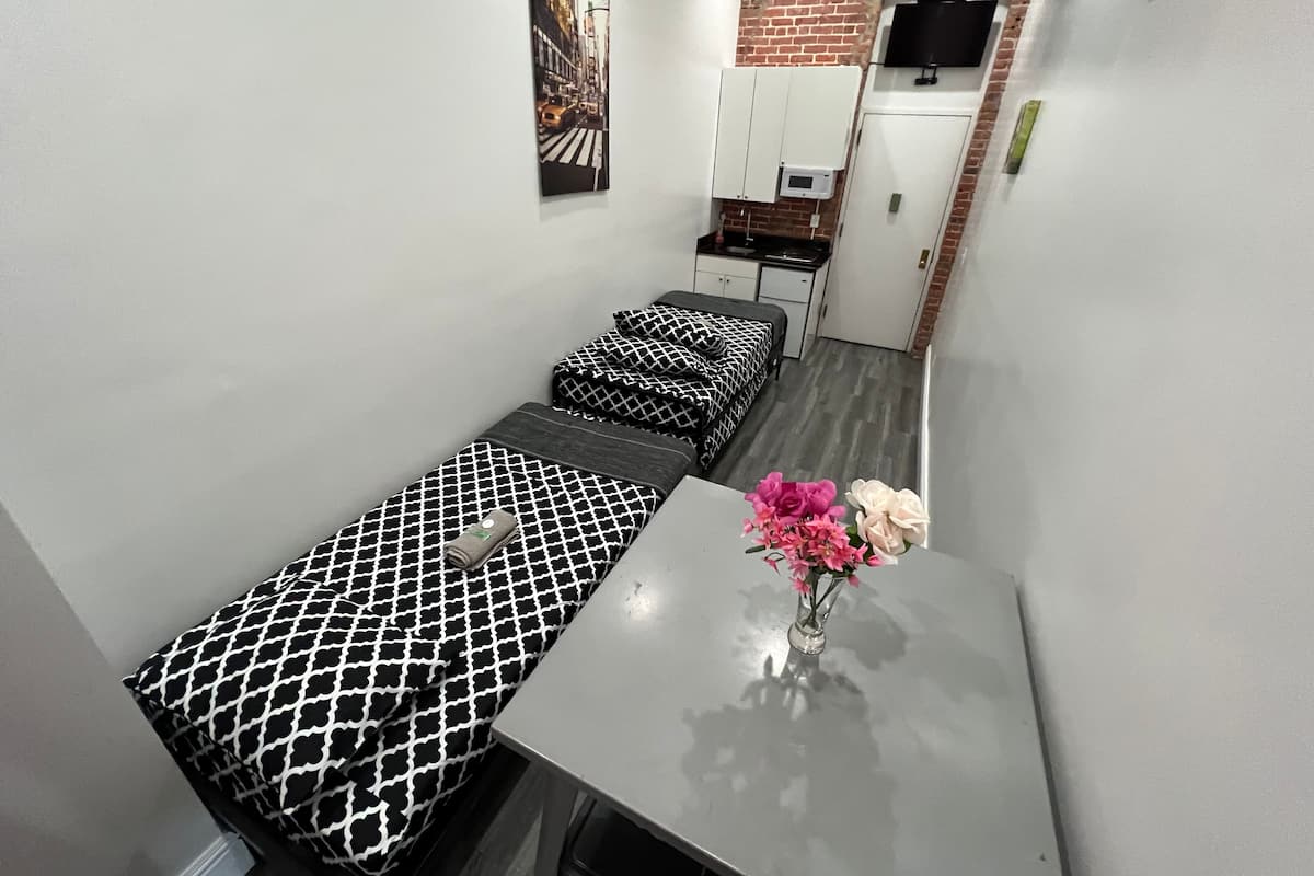 Una habitación pequeña y funcional con dos camas individuales, un kitchenette y un televisor montado en una pared de ladrillo.