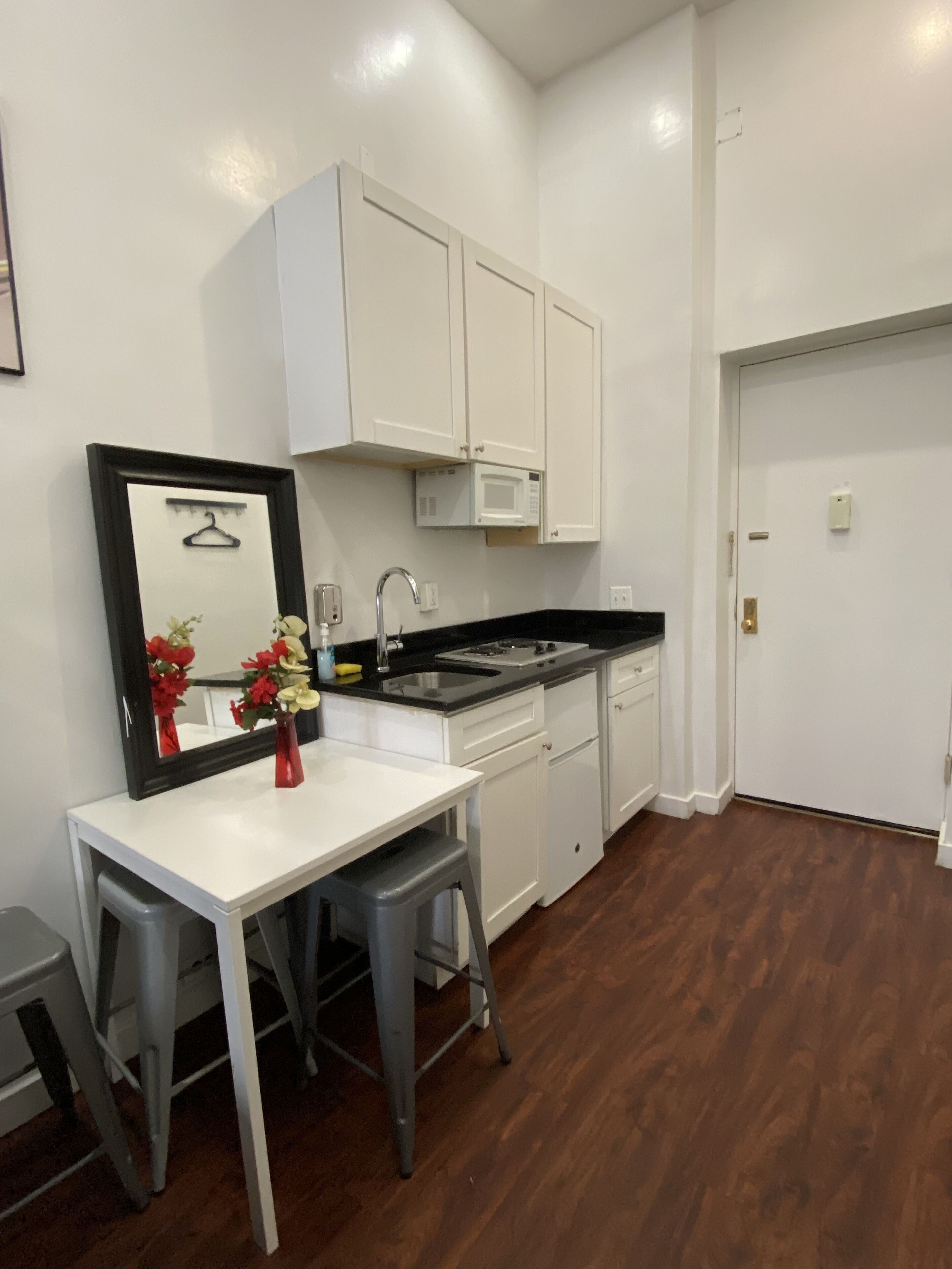Cocina compacta con armarios blancos, microondas y lavamanos. Hay una pequeña mesa de comedor con dos taburetes.