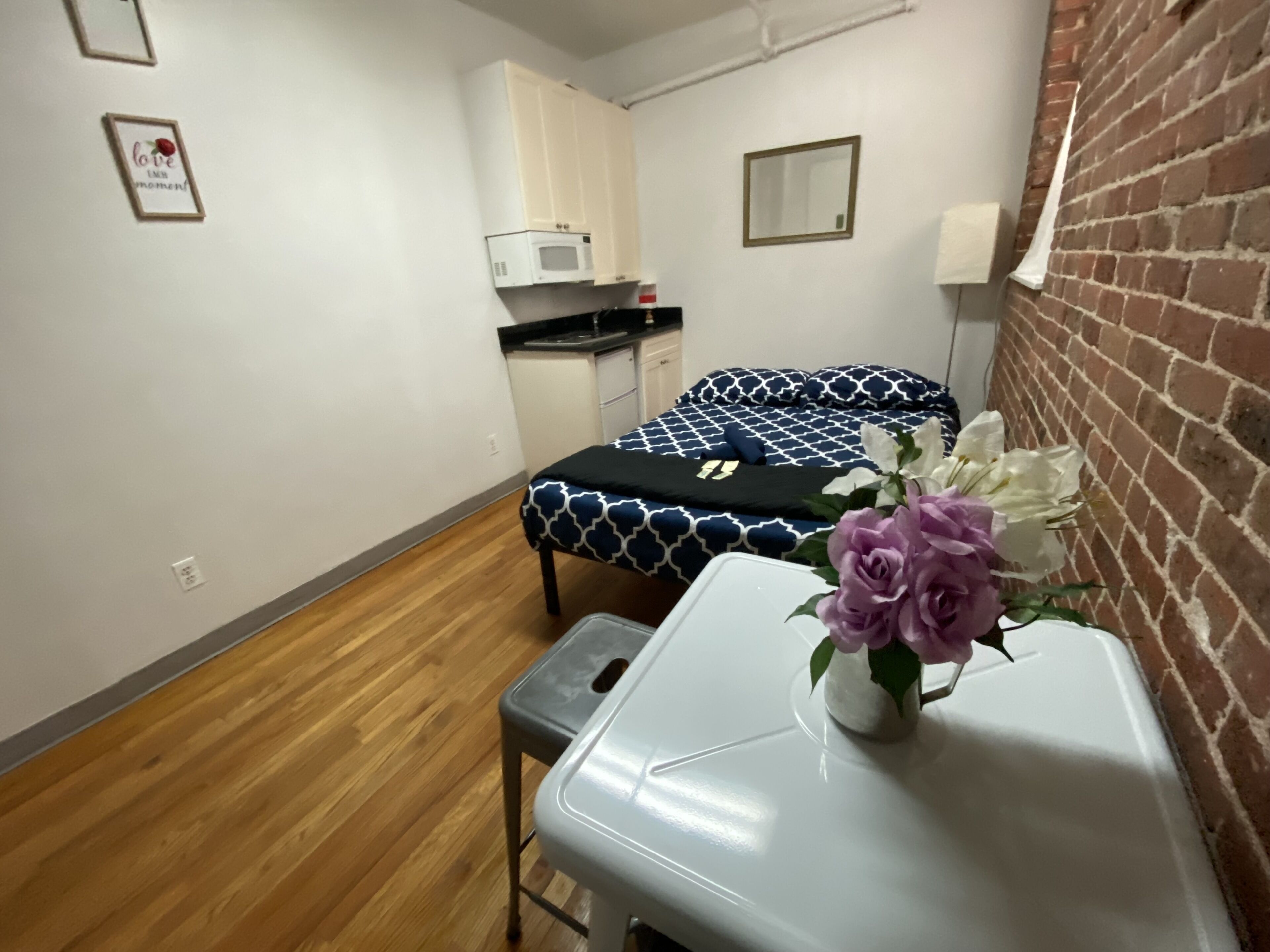 Une petite pièce, de taille modeste, comprenant un lit, une petite table avec un vase de fleurs, un four à micro-ondes et un mur en briques.