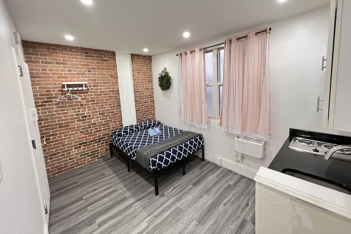 Una habitación con cama, pared de ladrillo, ventana con cortinas, radiador y kitchenette.