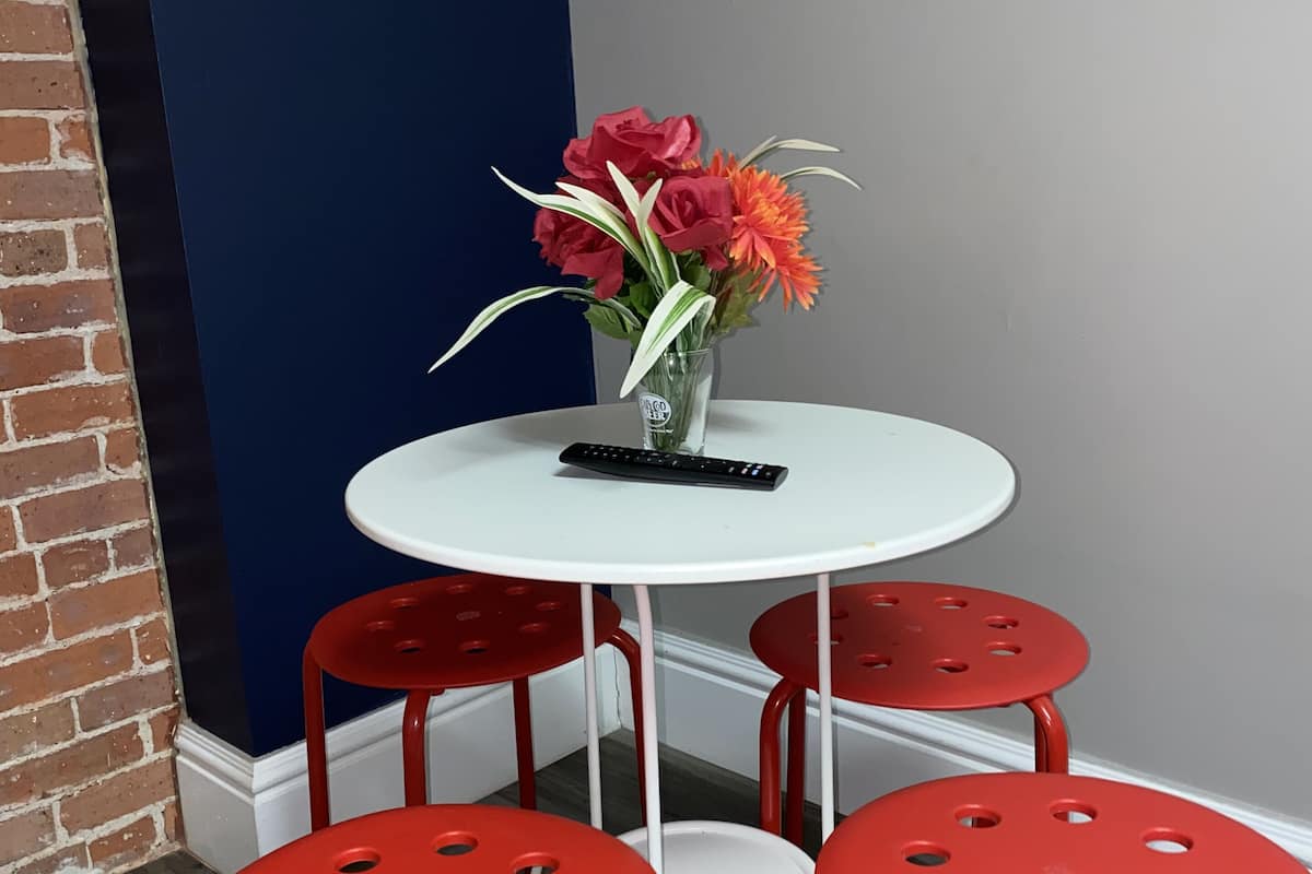 Un pequeño comedor con una mesa blanca y cuatro sillas rojas, un jarrón con flores y un control remoto sobre la mesa.