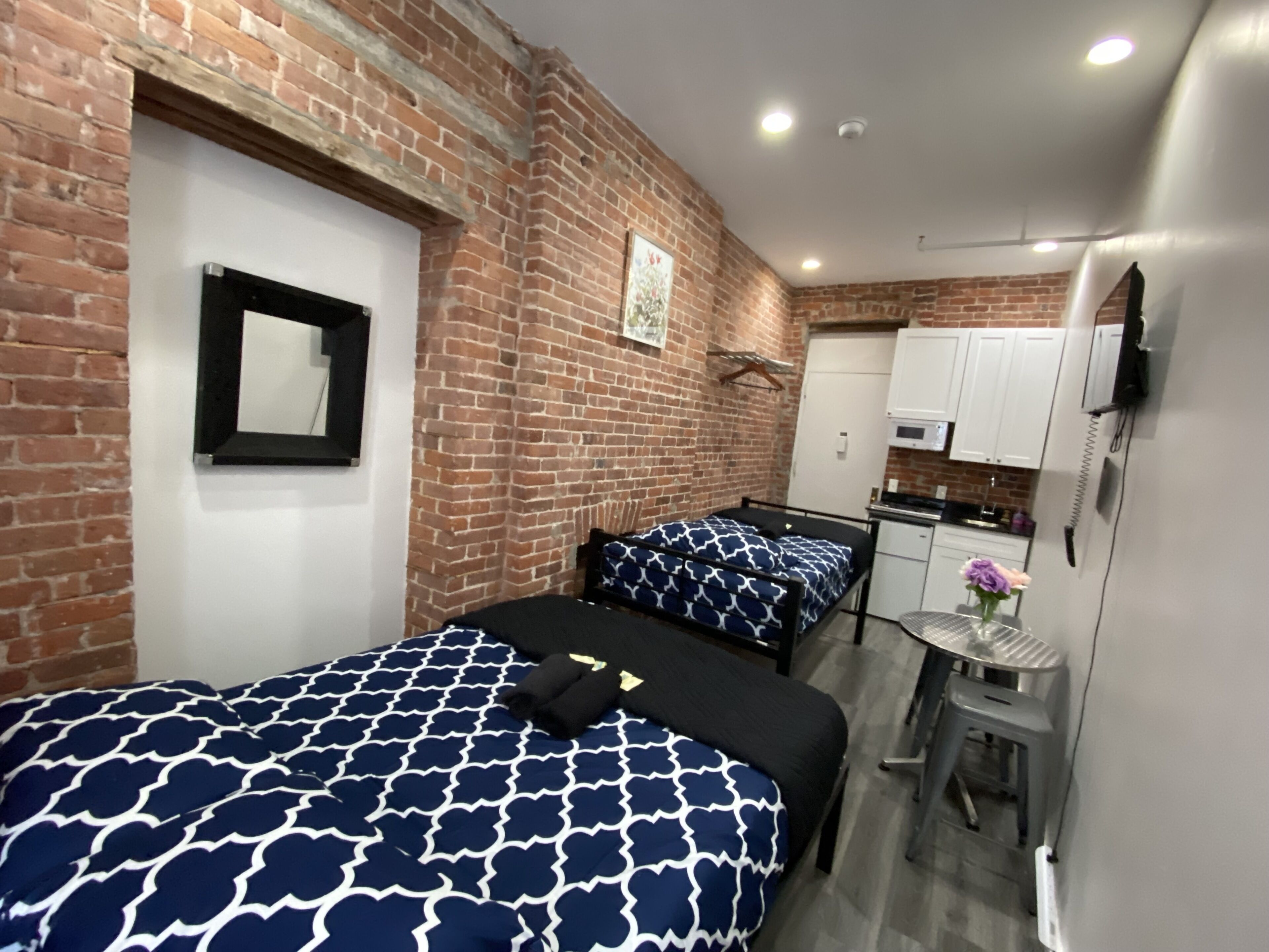 A compact room with two beds, a small kitchenette, and a TV mounted on the wall.