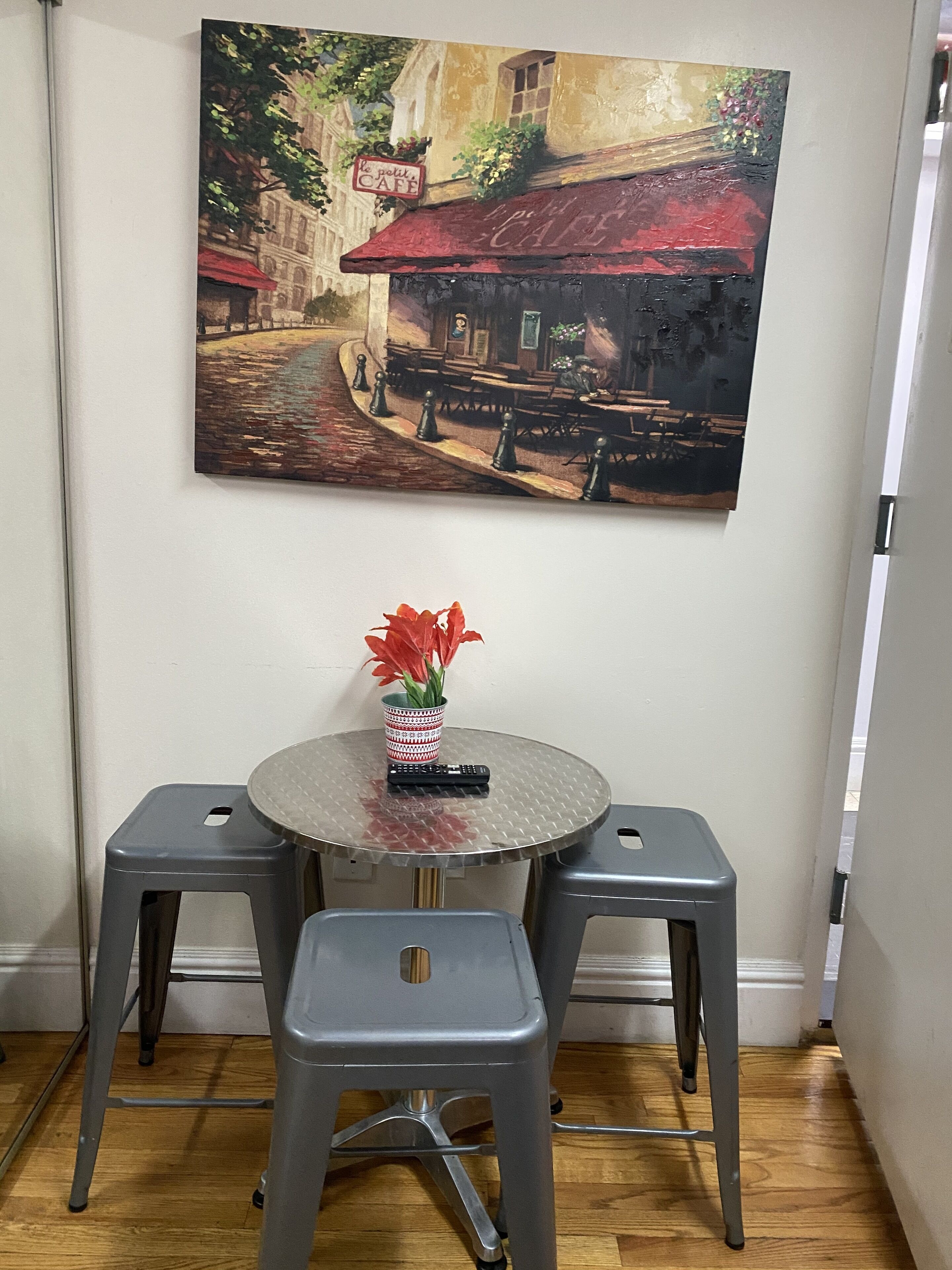 A dining area with a round table, two stools, a vase with red flowers, and a painting of a café on the wall.