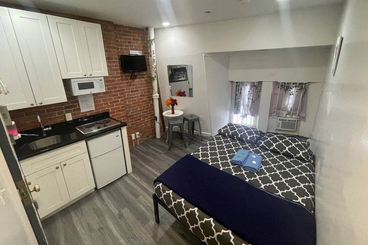 Habitación compacta con kitchenette, cama con ropa de cama estampada, una mesita con un florero y una ventana con cortinas.