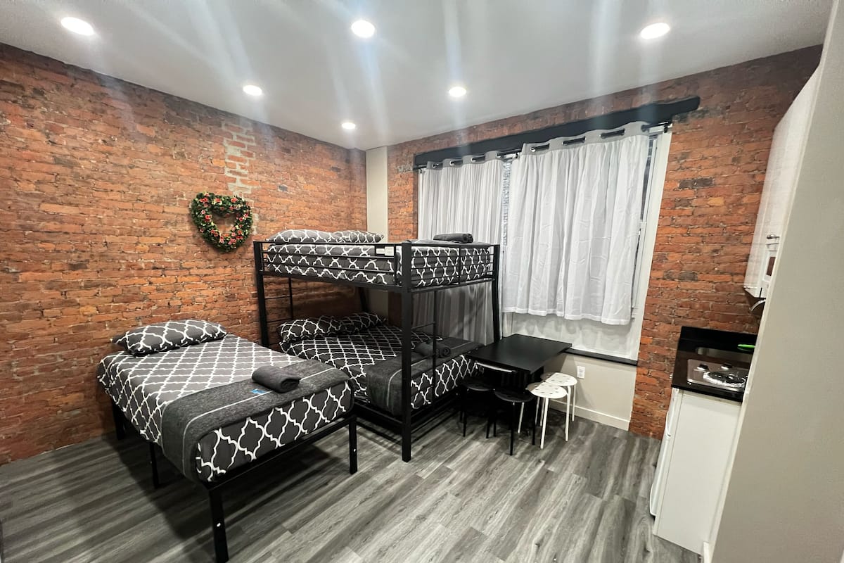 Una cama individual con ropa de cama estampada, una mesita con taburetes, un armario blanco y una pared de ladrillo con un adorno.