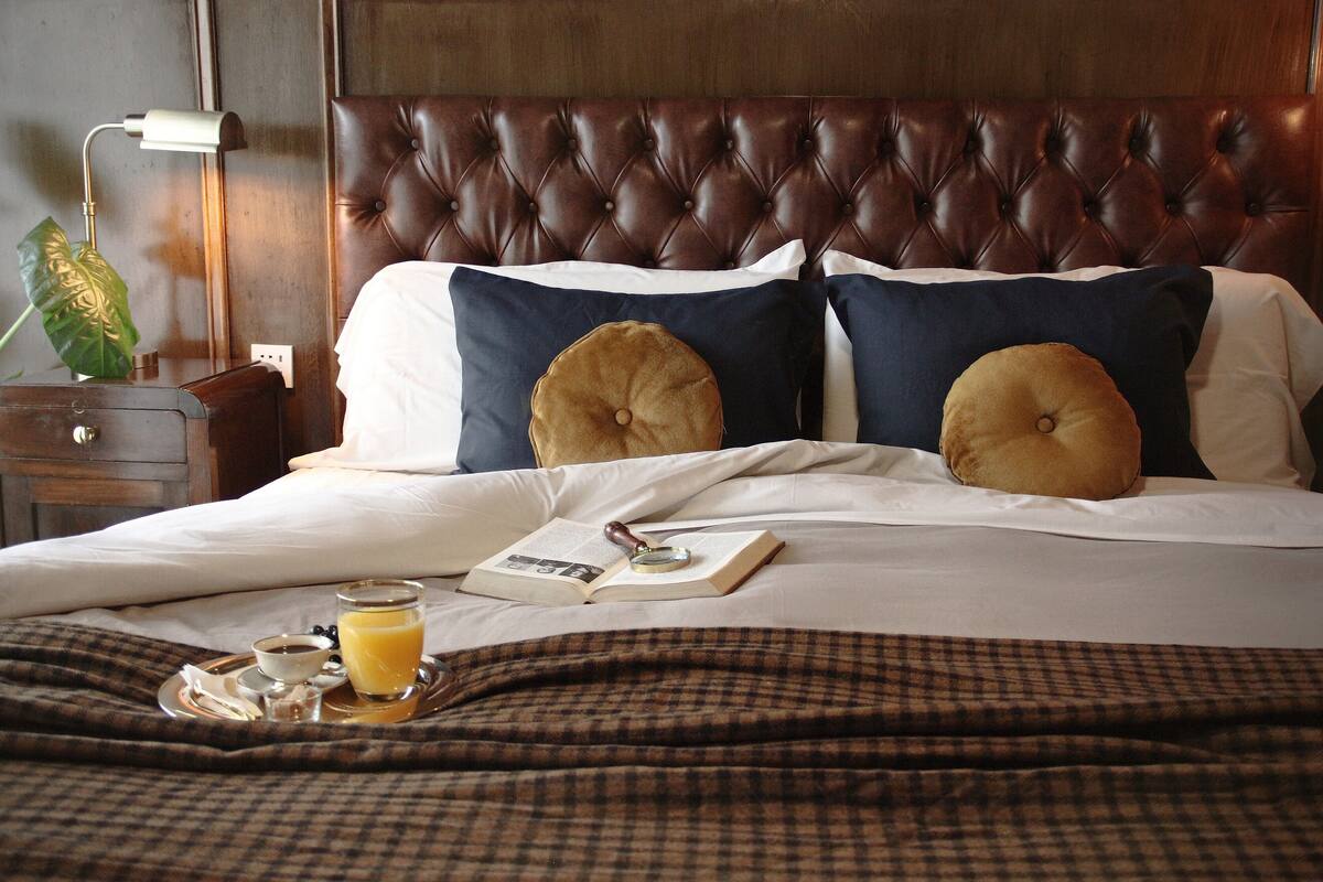 A neatly made bed with a checkered blanket, a bedside table with a lamp, and a tray with a book, glasses, and a glass of orange juice.