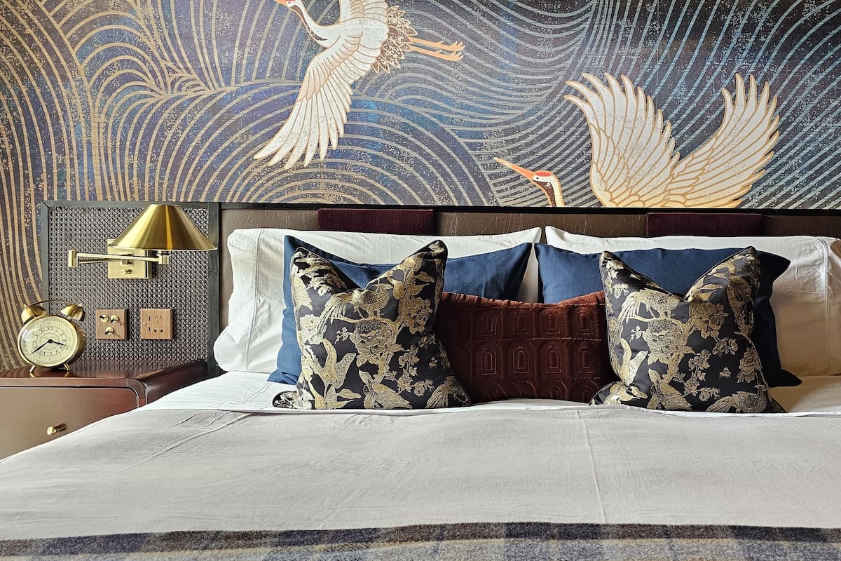 A bed with a white comforter and two decorative pillows, a wall clock, and a small table with a lamp and a plant.