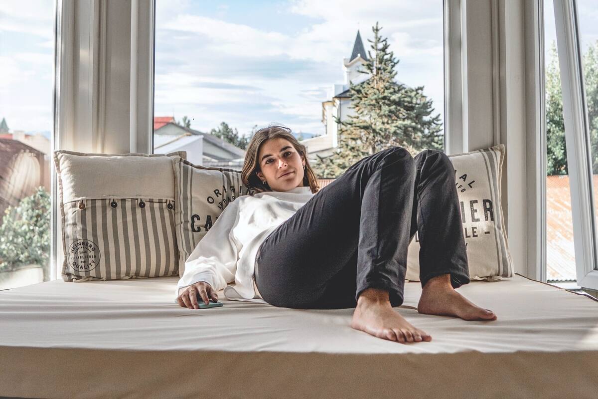 A person lying on a bed with a window view of a building and a tree.