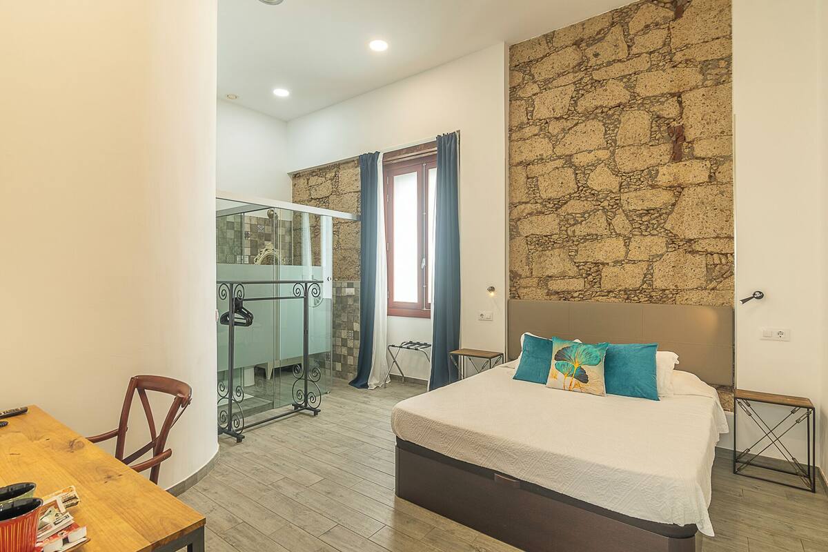A bedroom with a large bed, a wooden desk, a chair, a mirror, and a stone wall.