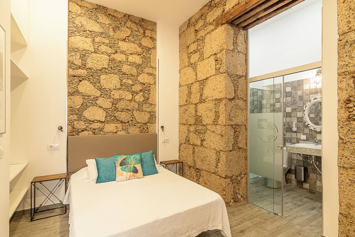 A bedroom with a stone wall, a bed with a white bedspread and teal pillows, a small bedside table, and a glass door leading to a bathroom with a tiled shower area.