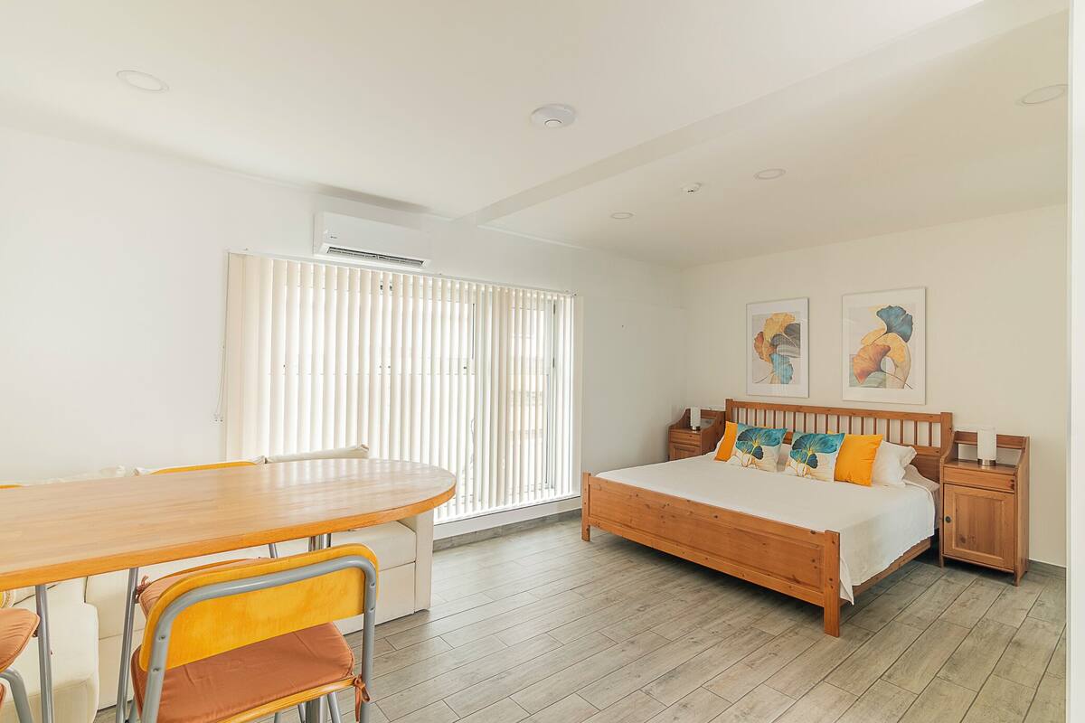 A bedroom with a wooden bed, a bedside table, a nightstand, a window with blinds, and a dining area with a table and chairs.