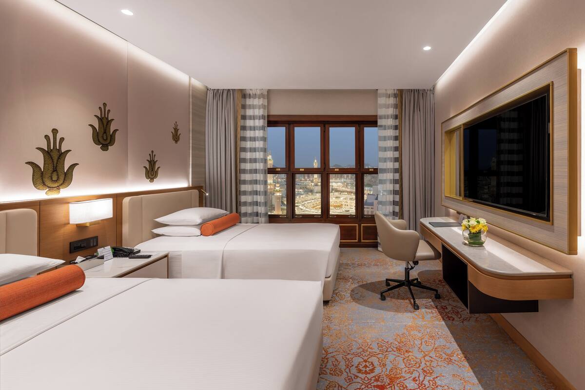 A hotel room with two beds, a desk, a TV, and a large window with a city view.