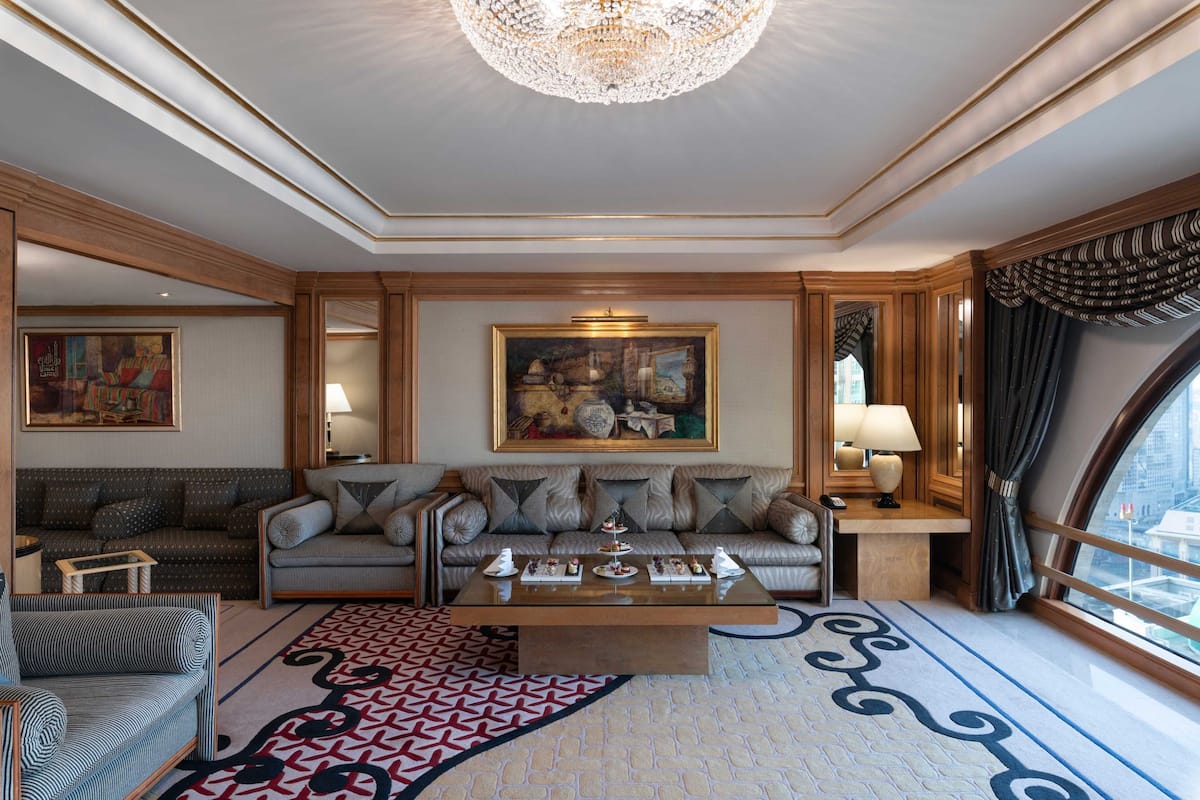 A luxurious living room with a large sofa set, a coffee table with decorative items, a chandelier, and a painting on the wall.