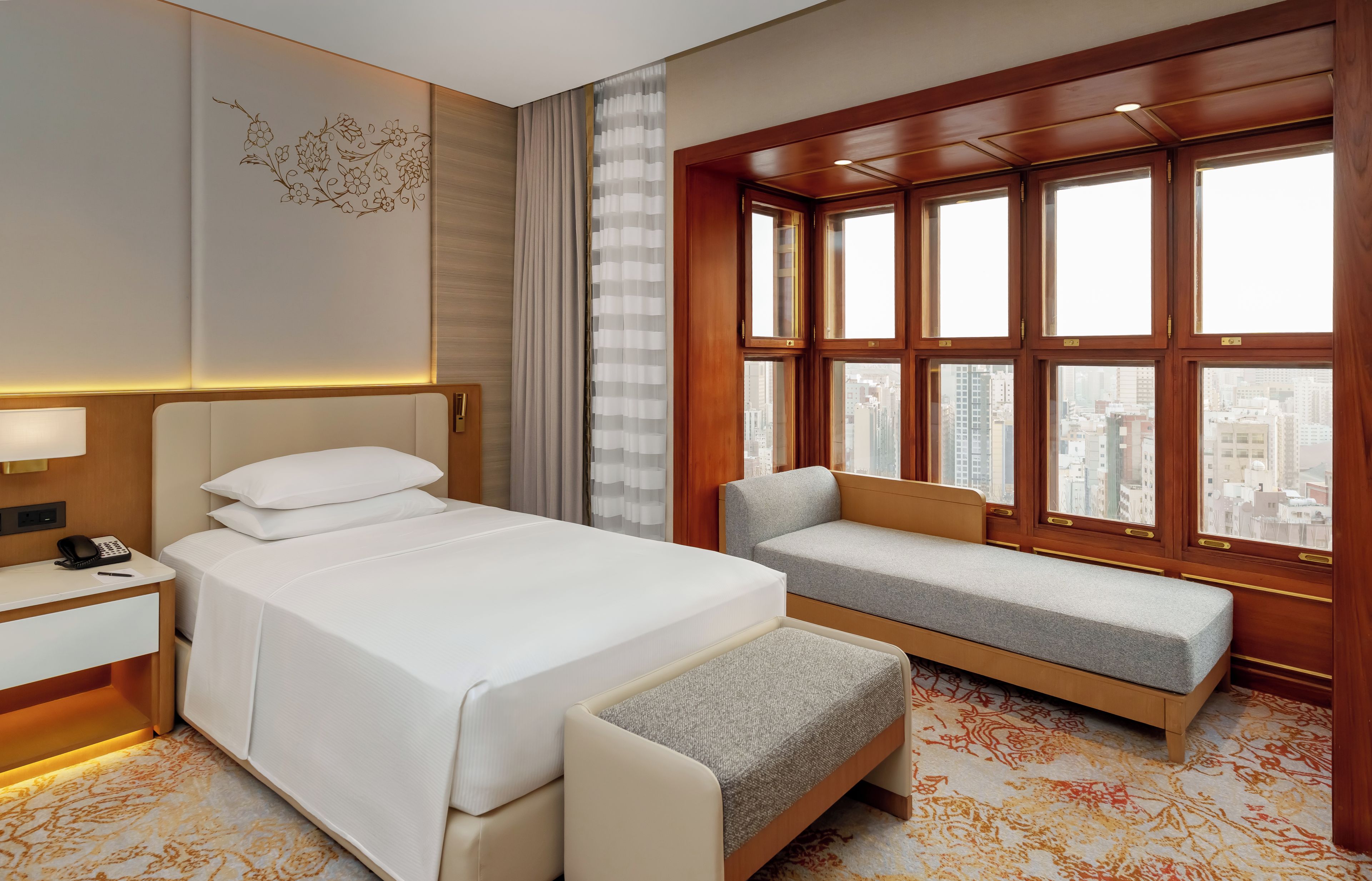 A hotel room with a bed, bedside table, lamp, and a view of the city through large windows.