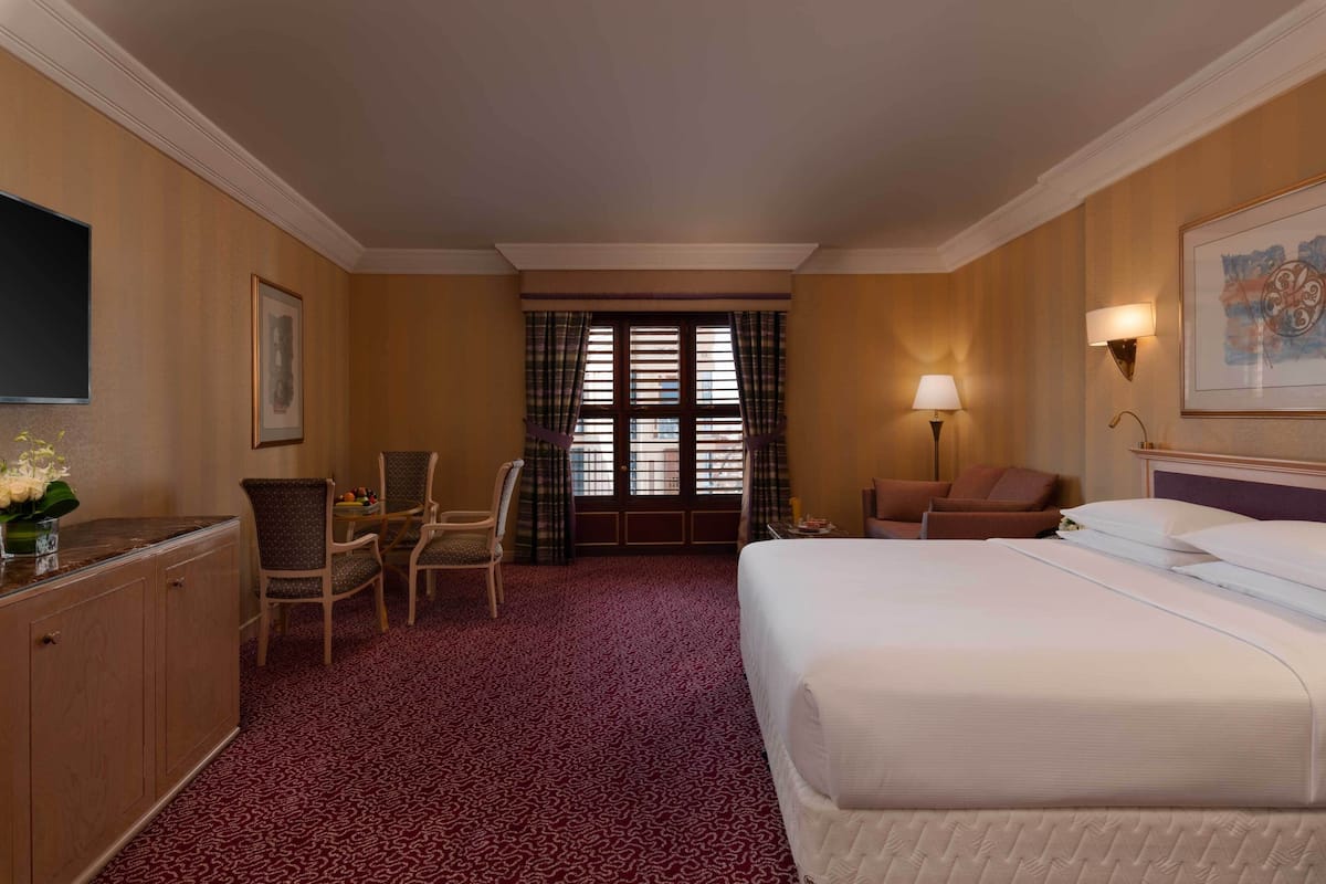 A hotel room with a large bed, a desk with a chair, a small table with a lamp, and a window with curtains.