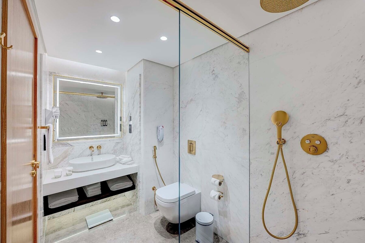 Un baño moderno con cabina de ducha de vidrio, un lavamanos blanco con detalles dorados y pared de mármol.