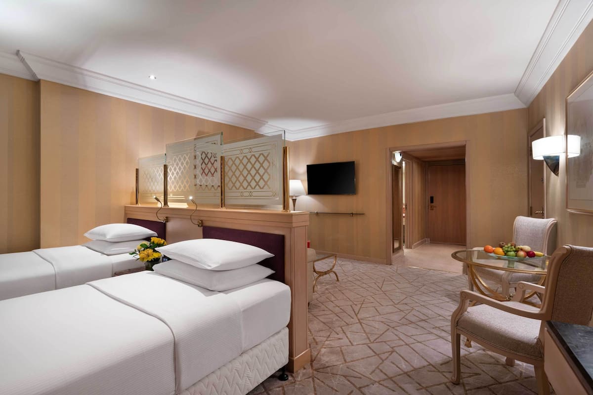 A hotel room with two beds, a sitting area with a table and chairs, a TV mounted on the wall, and a door leading to another room.