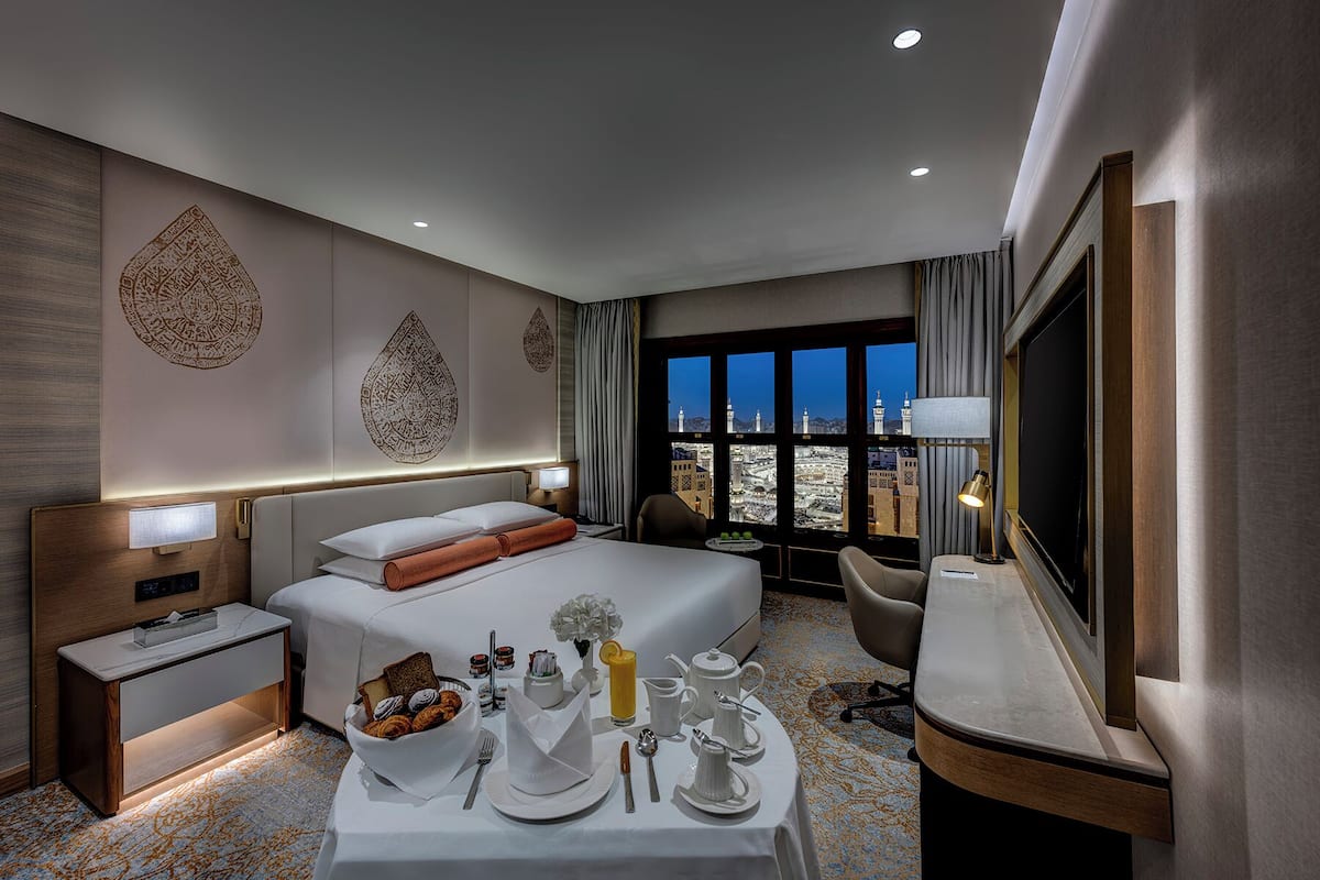 A modern hotel room with a bed, bedside table, desk, and a large window with a city view.