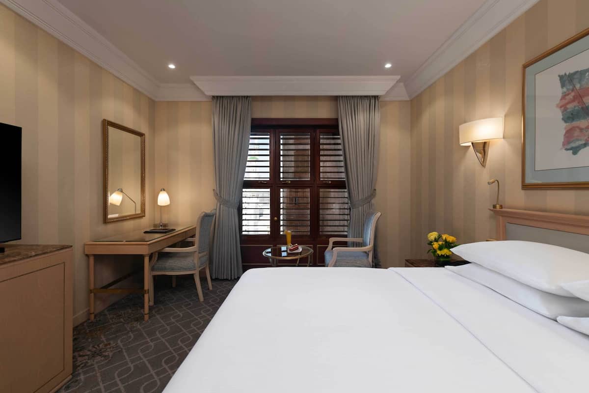 A hotel room with a bed, a desk, a chair, a small table, a TV, and a window with curtains.