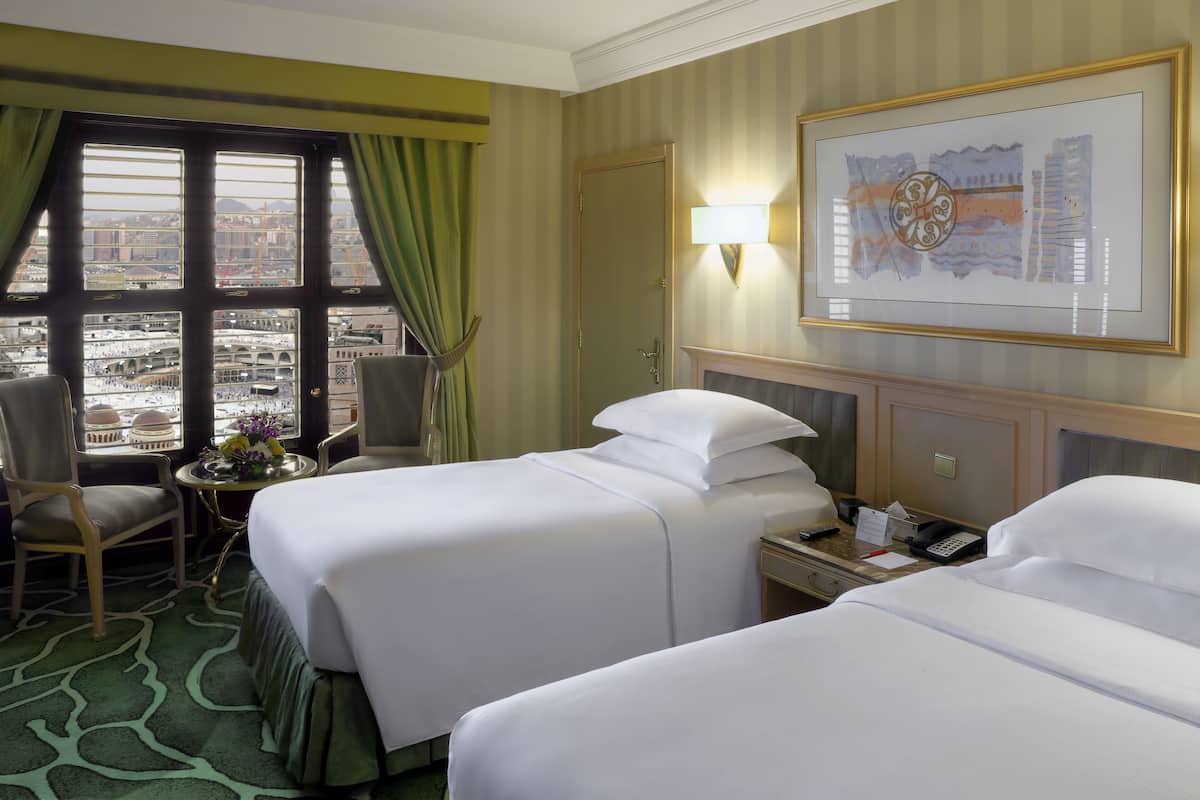 A hotel room with two beds, a desk, a chair, a window with curtains, a painting on the wall, and a view of the cityscape.
