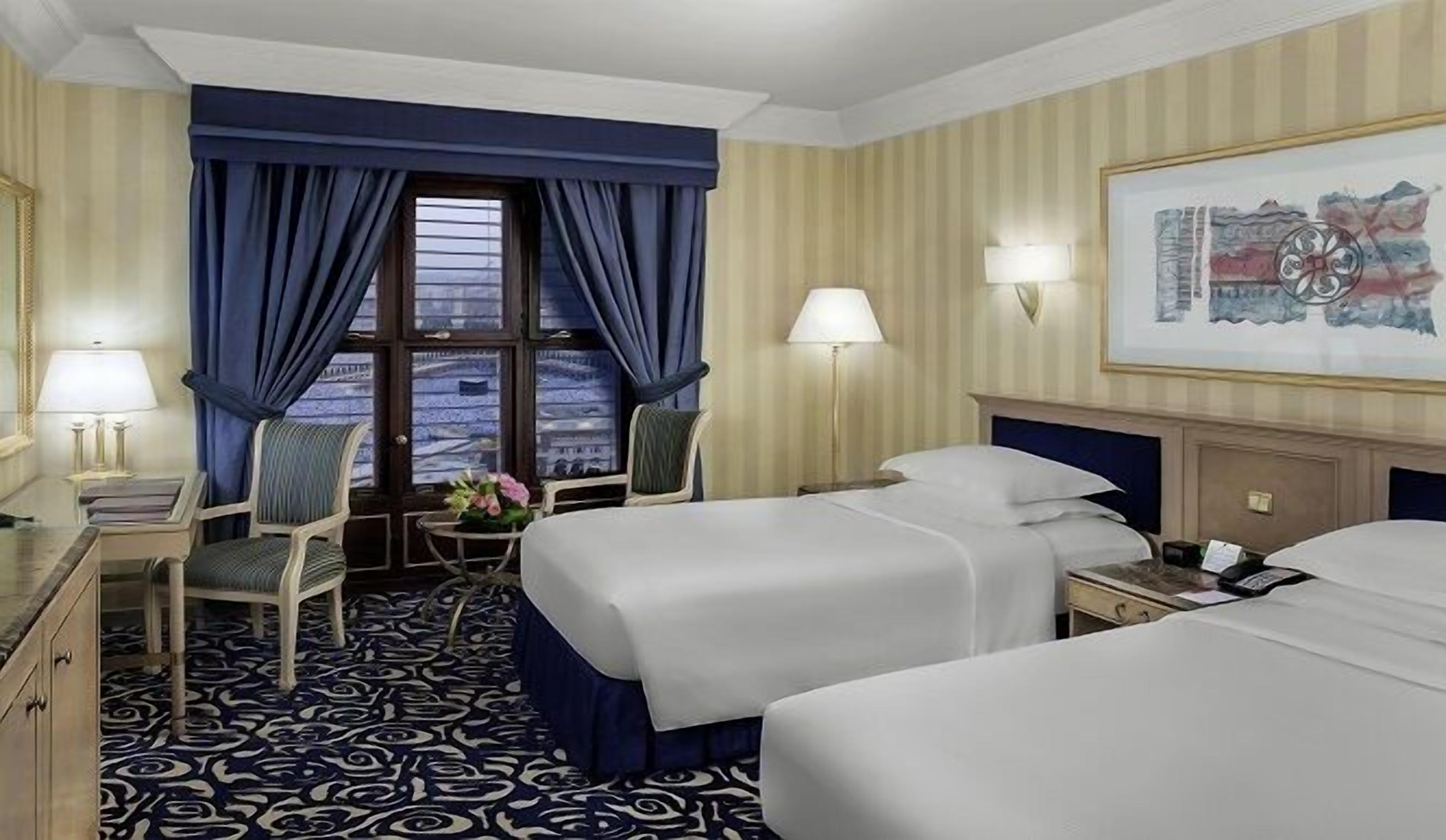 Ein Hotelzimmer mit zwei Betten, einem Schreibtisch mit Lampe, einem Sessel, einem Fenster mit Vorhängen und einem Bild an der Wand.
