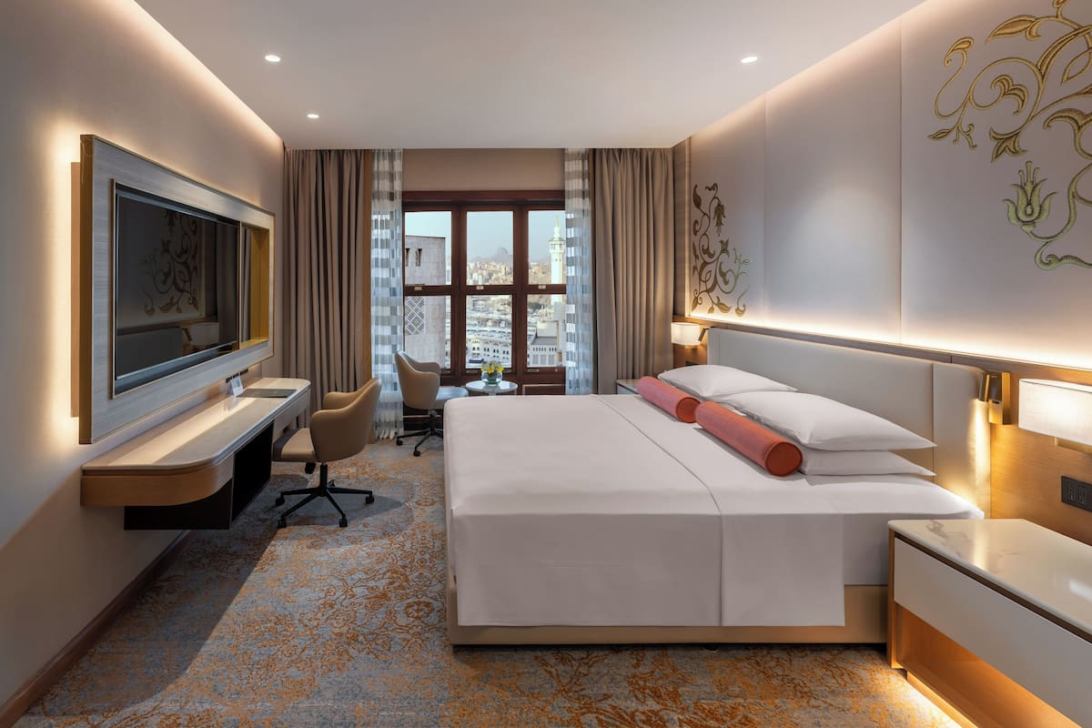 A modern hotel room with a large bed, a desk, a chair, a TV, and a view of the cityscape through the window.