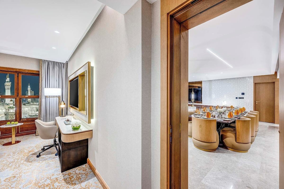 A modern hotel room with a large window, a dining area with wooden chairs, and a mirror with a light.