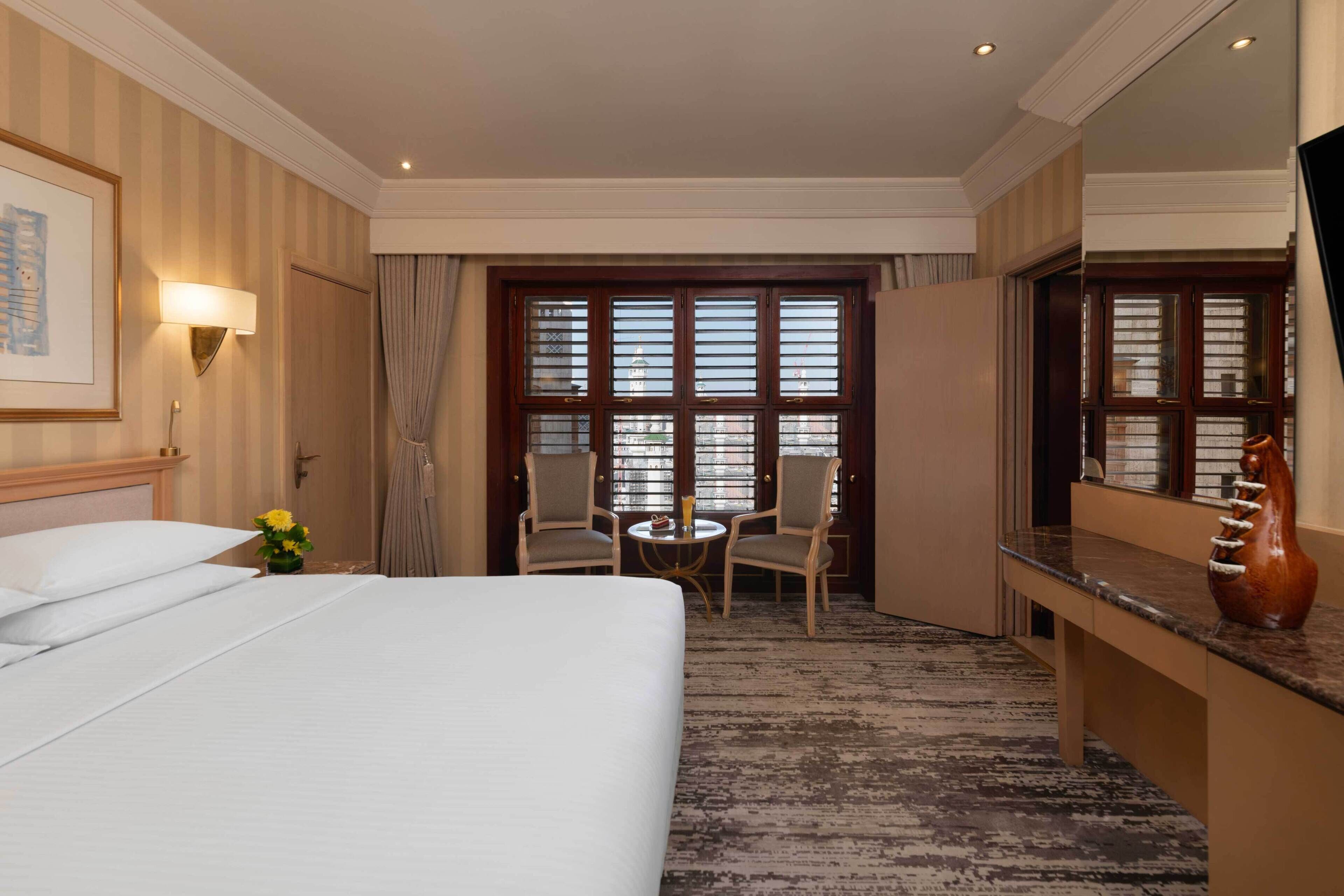 Habitación de hotel con una cama grande, un escritorio, una silla, una mesita y un ventanal con persianas.