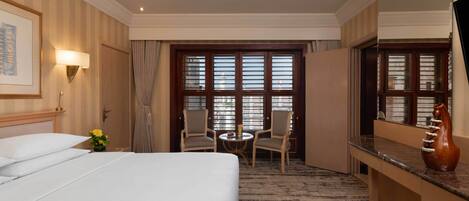 Habitación de hotel con una cama grande, un escritorio, una silla, una mesita y un ventanal con persianas.