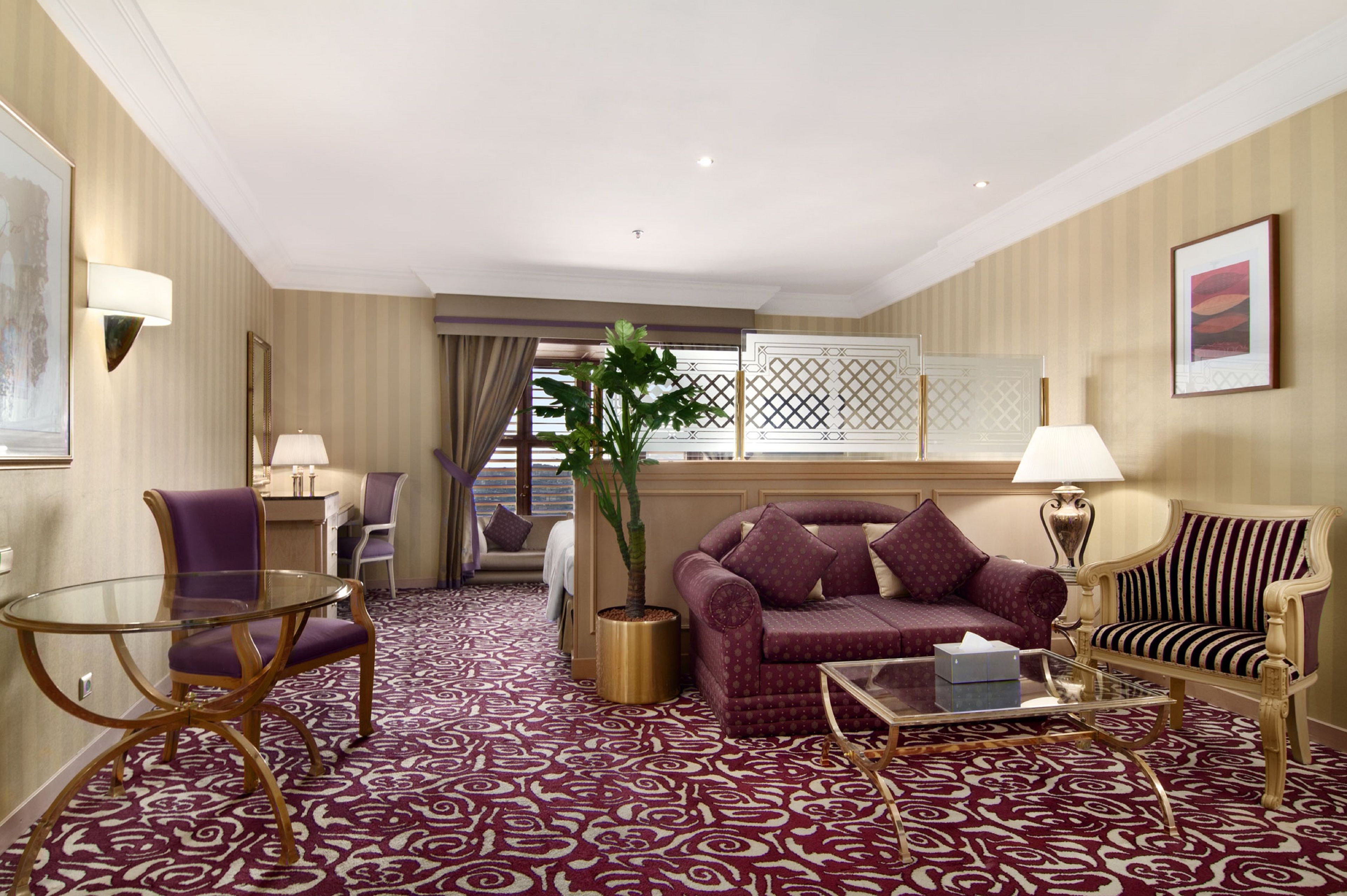 Habitación de hotel con alfombra estampada, un sofá, una silla, una mesa de centro de vidrio, una lámpara y una planta en maceta.
