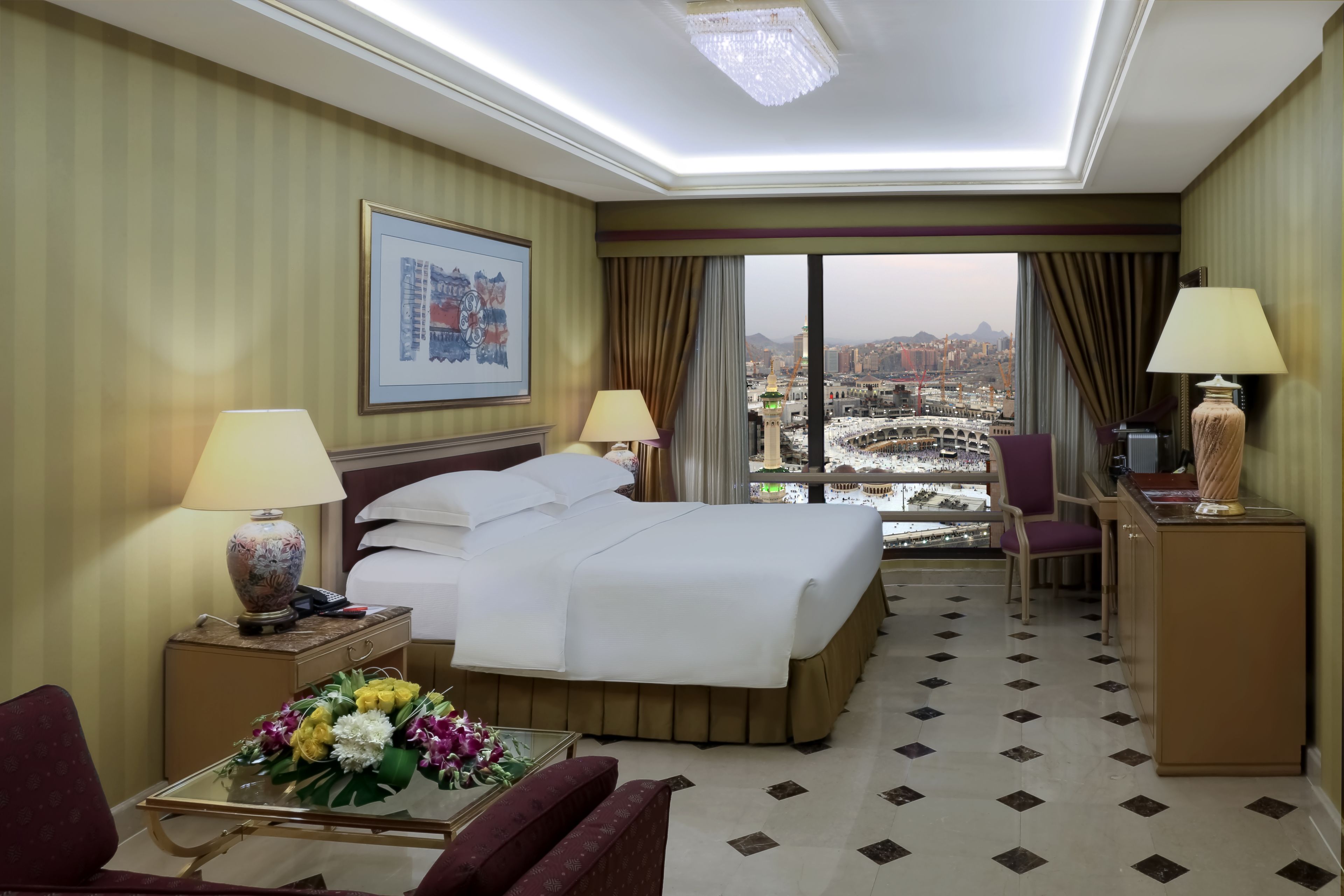 A hotel room with a large bed, two bedside lamps, a desk with a lamp, a chair, a small table with flowers, and a view of a cityscape through large windows.