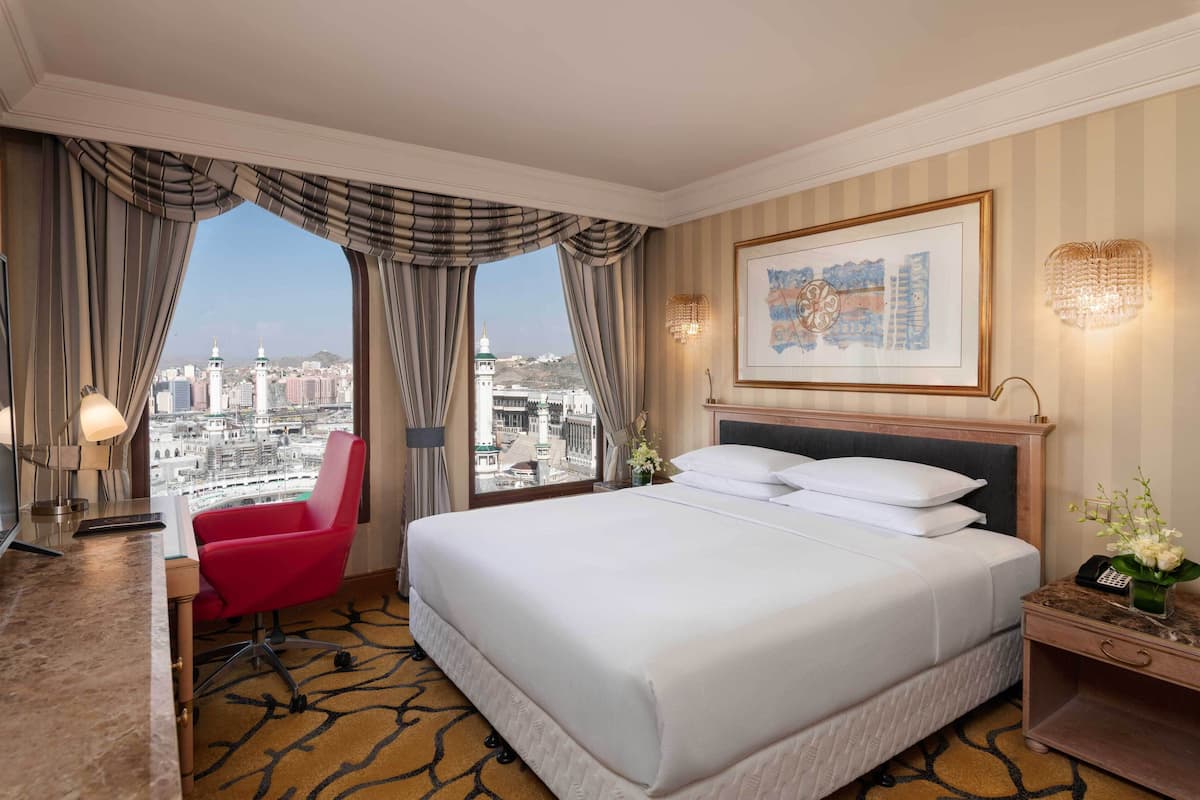 A hotel room with a large bed, a desk, a red chair, and a view of the cityscape through large windows.