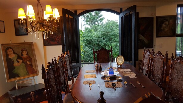 Una sala da pranzo con un grande tavolo imbandito per sei persone, circondato da sedie elaborate. Sopra, un lampadario e, sulla parete, un dipinto.