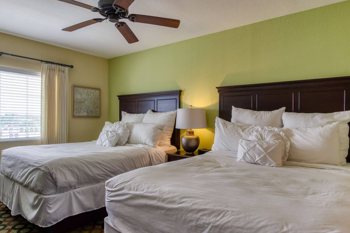 A hotel room with two beds, a ceiling fan, a nightstand with a lamp, and a window with curtains.