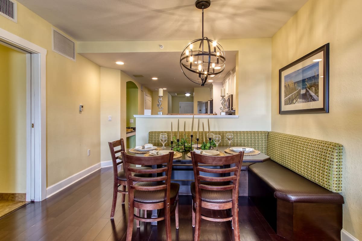 A dining area with a round table set for six, surrounded by wooden chairs, a bench, and a chandelier.
