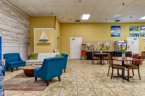 A hotel lobby with a yellow wall, a white door, blue armchairs, a wooden coffee table, a dining area with a wooden table and chairs, and a sailboat decoration on the wall. - Harbourside Resort, a New Hotel Collection Property   (Indian Rocks Beach)