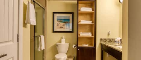 A bathroom with a toilet, a glass shower enclosure, a sink with a granite countertop, and a shelf with folded towels.